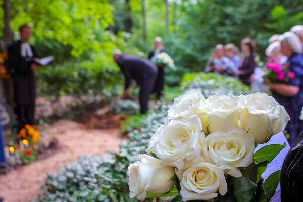 Roses in cemetery with people in the background. Funeral in cemetery;  White roses in cemetery. Prayer at the tomb. Funeral ceremony. White roses at funeral near the grave.