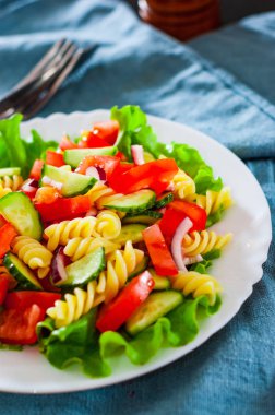 domates, marul, soğan ve masada beyaz tabak içinde salatalık sebze makarna salatası