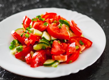 siyah arka plan üzerine beyaz tabakta domates, salatalık ve yeşil soğan ile taze sebze salatası.