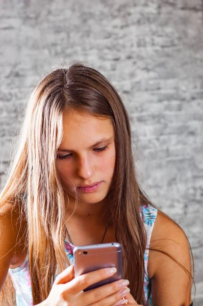 灰色の壁の背景に携帯電話を使用して長い髪の 代の若いブルネットの少女の肖像画 — ストック写真