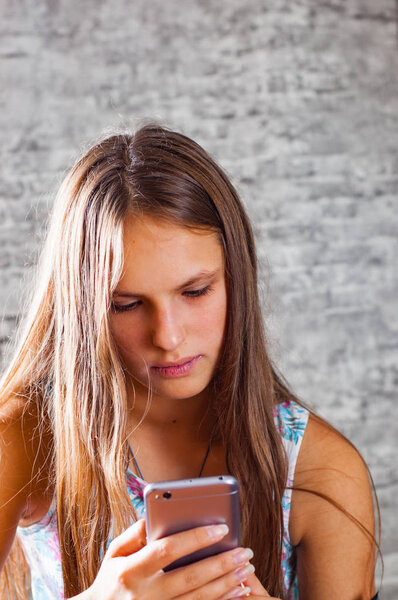 portrait of young teenager brunette girl with long hair using mobile phone on gray wall background