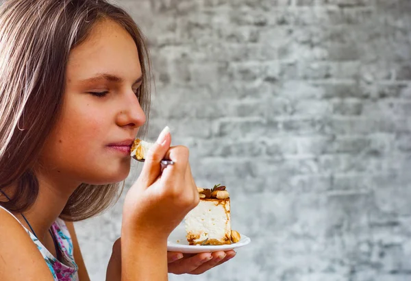 灰色の壁の背景にケーキ デザートを食べて長い髪の 代の若いブルネットの少女の肖像画 — ストック写真