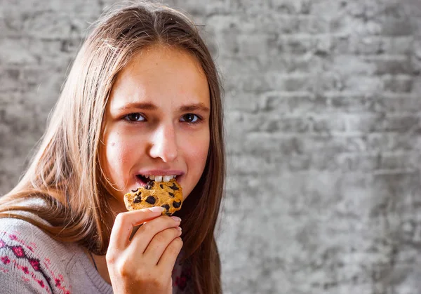 灰色の壁の背景にクッキーやビスケットを食べる長い髪の 代の若いブルネットの少女の肖像画 — ストック写真