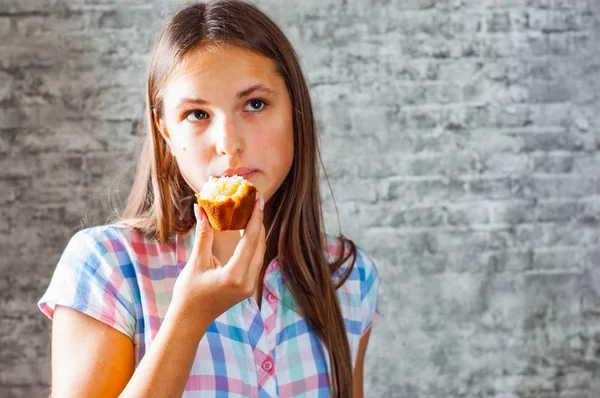 灰色の壁の背景にマフィン ケーキを食べる長い髪の 代の若いブルネットの少女の肖像画 — ストック写真