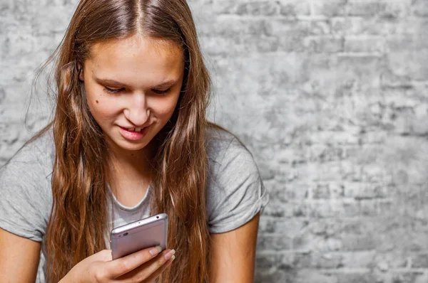 灰色の壁の背景に携帯電話を使用して長い髪の 代の若いブルネットの少女の肖像画 — ストック写真