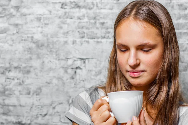 灰色の壁背景でコーヒーまたは紅茶のカップを保持している長い髪の 代の若いブルネットの少女の肖像画 — ストック写真