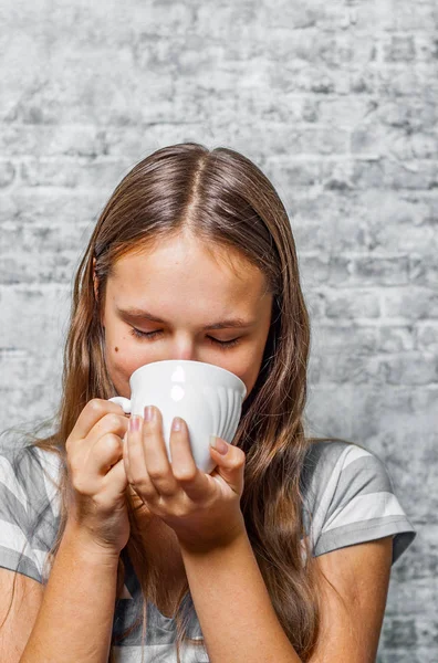 灰色の壁背景でコーヒーまたは紅茶のカップを保持している長い髪の 代の若いブルネットの少女の肖像画 — ストック写真