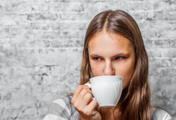 灰色の壁背景でコーヒーまたは紅茶のカップを保持している長い髪の 代の若いブルネットの少女の肖像画 — ストック写真