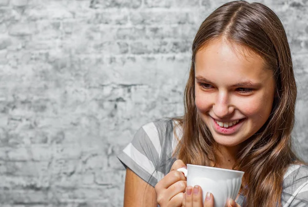 灰色の壁背景でコーヒーまたは紅茶のカップを保持している長い髪の 代の若いブルネットの少女の肖像画 — ストック写真