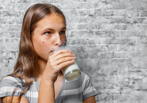 灰色の壁の背景に牛乳のガラスを保持している長い髪の若い 代のブルネットの少女の肖像画 — ストック写真