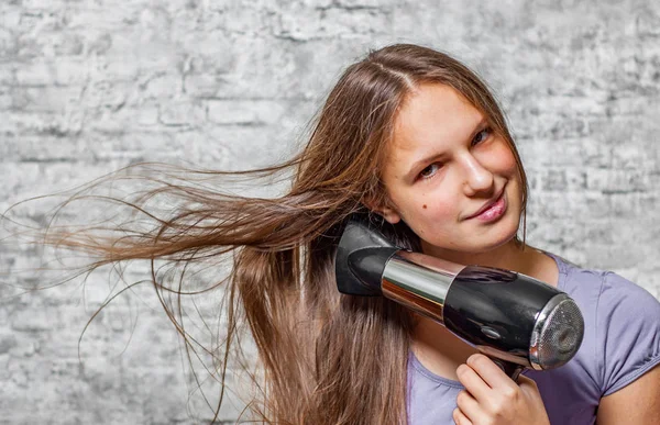 長い髪の若い 代のブルネットの少女の肖像画は灰色の壁の背景に電気ドライヤーで髪を乾燥します — ストック写真