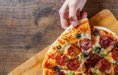 Kadın el Pizza bir dilim Mozzarella peyniri, sucuk, domates, biber, zeytin, salam ile alır. Ahşap masa arka plan üzerinde İtalyan pizza