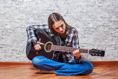 katta oturan ve gri duvar arka plan üzerinde siyah akustik gitar çalmaya uzun saçlı Genç Genç esmer kız