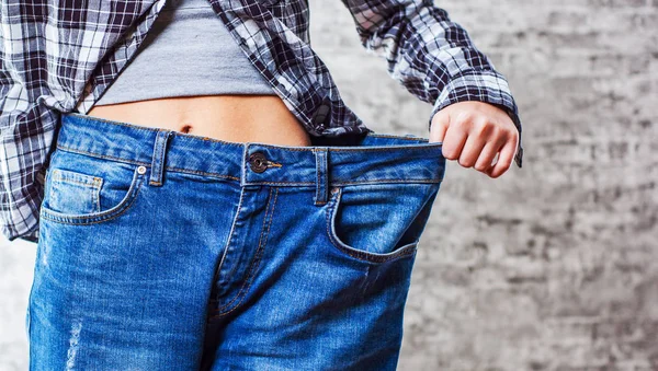 Diet concept and weight loss. Woman shows her weight loss by wearing an old jeans on gray wall background