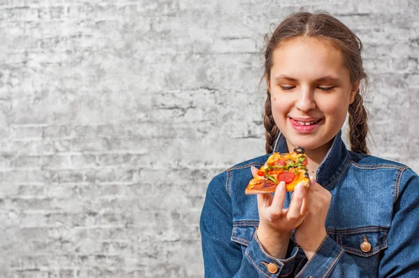 コピー スペースを持つ灰色の壁背景にピザのスライスを食べる長い髪の若いティーネー ジャーのブルネットの少女の肖像画 — ストック写真