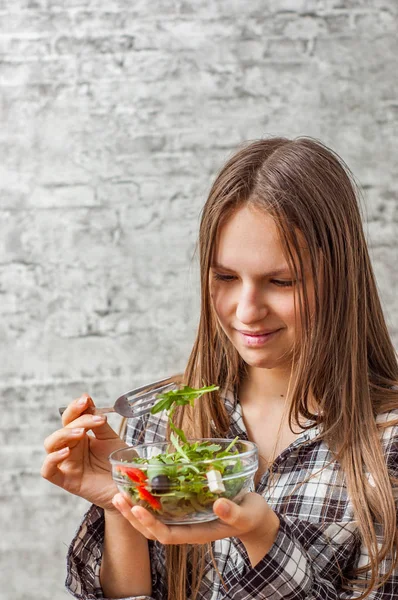 灰色の壁の背景に緑の野菜サラダを食べて長い髪の 代の若いブルネットの少女の肖像画 — ストック写真