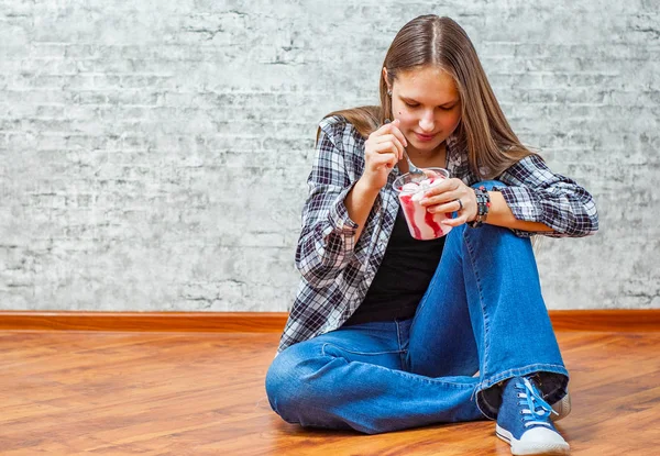 灰色の壁の背景に床に座ってアイスクリームを食べて長い髪の 代の若いブルネットの少女の肖像画 — ストック写真