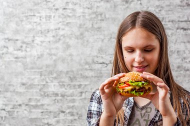 Hamburger yemek uzun saçlı genç genç esmer kız portresi. Kız gri duvar arka plan üzerinde hızlı yemek yemeye çalışıyor