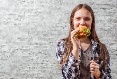 Hamburger yemek uzun saçlı genç genç esmer kız portresi. Kız gri duvar arka plan üzerinde hızlı yemek yemeye çalışıyor