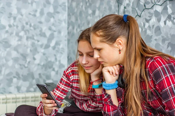 灰色の壁の背景に自宅でソファーに座っている間 携帯電話を使っている2人の若い十代の少女 — ストック写真