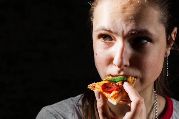 portrait of young teenager brunette girl with long hair eating slice of pizza on dark black wall background with copy space.