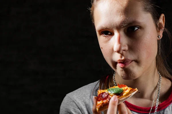 portrait of young teenager brunette girl with long hair eating slice of pizza on dark black wall background with copy space.