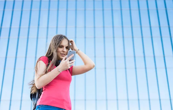 長い髪のサンゴのTシャツで若いティーンエイジャーブルネットの女の子の屋外の肖像画 スマートフォンを見て街の女の子 コピースペースを持つ背景にガラスの建物 — ストック写真