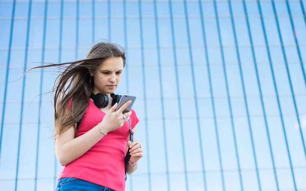 長い髪のサンゴのTシャツで若いティーンエイジャーブルネットの女の子の屋外の肖像画 スマートフォンを見て街の女の子 コピースペースを持つ背景にガラスの建物 — ストック写真