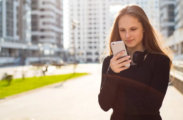 長い髪を持つ若いティーンエイジャーブルネットの女の子の屋外の肖像画 晴れた日に通りでスマートフォンを見て黒いドレスの街の女の子 — ストック写真