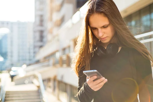 長い髪を持つ若いティーンエイジャーブルネットの女の子の屋外の肖像画 晴れた日に通りでスマートフォンを見て黒いドレスの街の女の子 — ストック写真