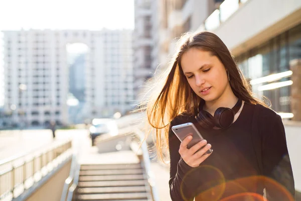 長い髪を持つ若いティーンエイジャーブルネットの女の子の屋外の肖像画 晴れた日に通りでスマートフォンを見て黒いドレスの街の女の子 — ストック写真