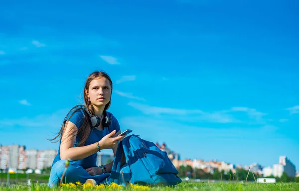 户外肖像的年轻的青少年黑发女孩与长发 女孩与智能手机和耳机坐在草地上 在公园的天空与复制空间 — 图库照片