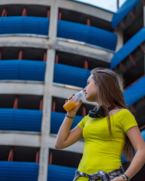 長い髪を持つ若いティーンエイジャーブルネットの女の子の屋外の肖像画 十代の少女は 都市の駐車場の背景に新鮮なオレンジジュースのボトルを飲みます — ストック写真