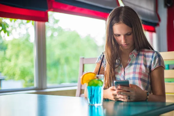 都会のカフェで屋内に座って長い髪を持つ若いティーンエイジャーブルネットの女の子の肖像画は 青いレモネードカクテルを飲み 彼女のスマートフォンを使用します — ストック写真