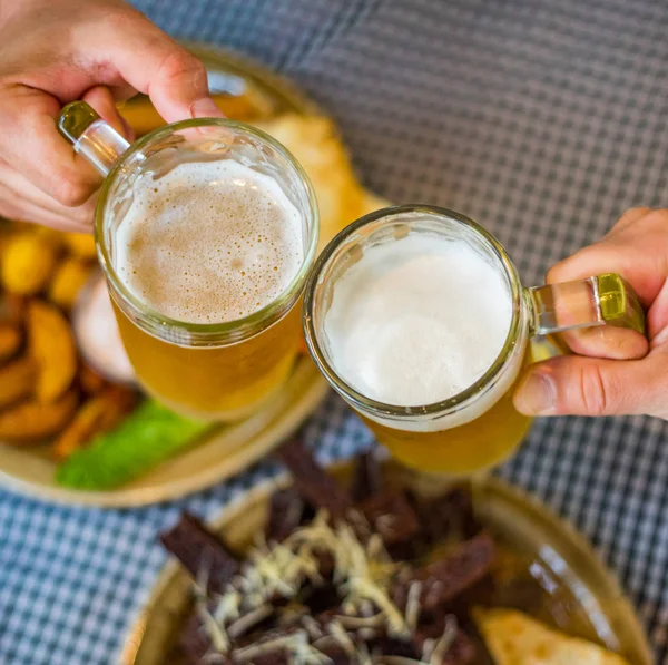Close-up view of a two glass of beer in hand. Beer glasses clinking in bar or pub on table with food background