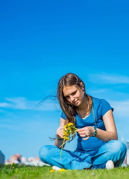 長い髪を持つ若いティーンエイジャーブルネットの女の子は草の上に座って コピースペースと空の背景に黄色のタンポポの花の花輪をリース — ストック写真