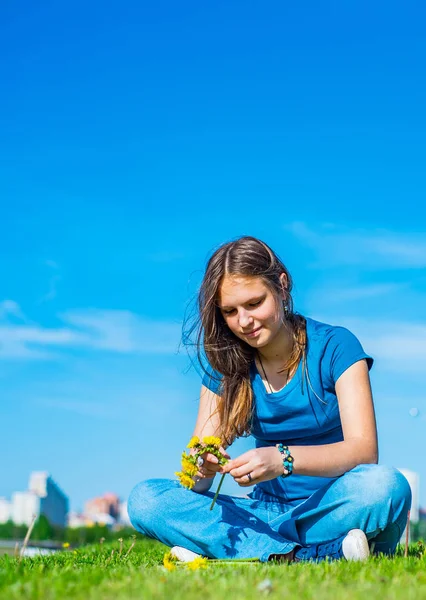 長い髪を持つ若いティーンエイジャーブルネットの女の子は草の上に座って コピースペースと空の背景に黄色のタンポポの花の花輪をリース — ストック写真