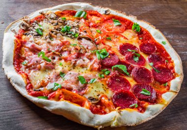 Mozzarella peynirli, jambonlu, domates soslu, salamlı, pastırmalı, mantarlı, biberli, baharatlı ve taze fesleğenli pizza. İtalyan pizzası ahşap masa arkasında