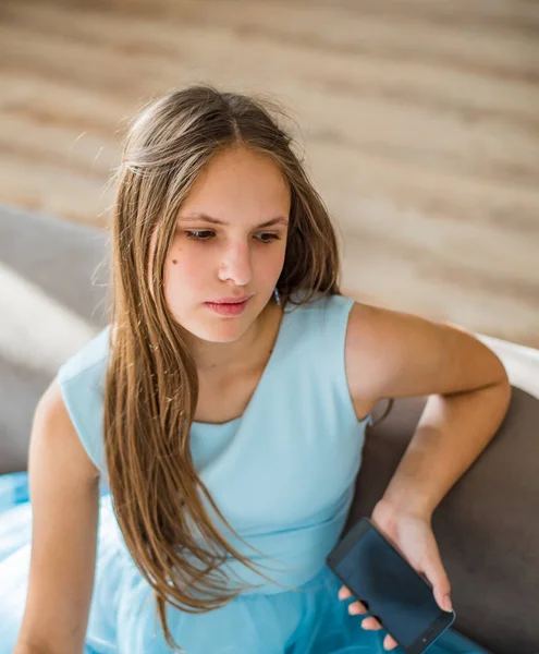 スマートフォンと青いドレスで自宅のソファに座って長い髪を持つ若いティーンエイジャーブルネットの女の子の肖像画 — ストック写真