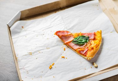 Mozzarella peyniri, jambon, pastırma, Domates sosu, biber, Baharatlar ve fesleğen ile Pizza son bir dilim. Ahşap masa arka plan üzerinde karton kutu içinde İtalyan pizza