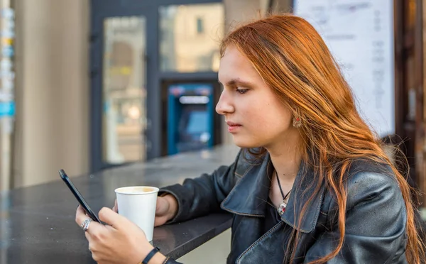 若いですティーン赤毛の女の子とともに長い髪でスマートフォンやカップのホットコーヒーで街のカフェテラス — ストック写真