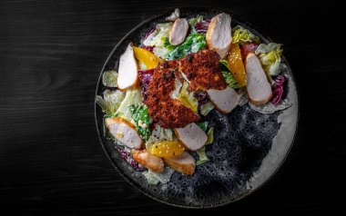 Tavuk, yumurta, mandalina ve sebzeli salata siyah ahşap masa arkasında.