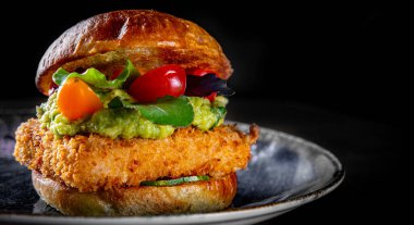 Peynirli balık burger, guacamole sosu ve siyah ahşap masa arka planında sebzeler.