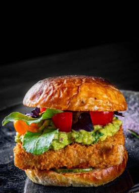Peynirli balık burger, guacamole sosu ve siyah ahşap masa arka planında sebzeler.