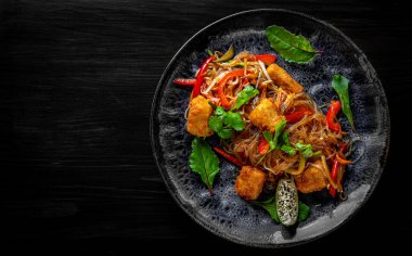 Lezzetli Asya eriştesi, kızarmış peynirli tofu ve sebzeli siyah ahşap masa arkaplanı.