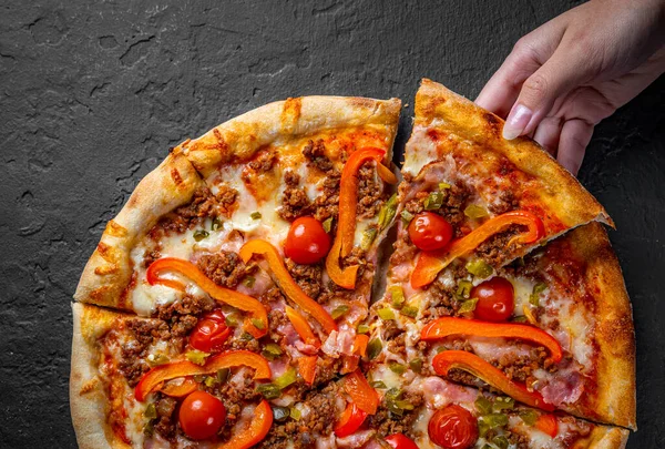 woman Hand takes a slice of Pizza with Mozzarella cheese, Bolognese sauce, minced meat, pepper, tomato, bacon and vegetables. Italian pizza on Dark grey black slate background