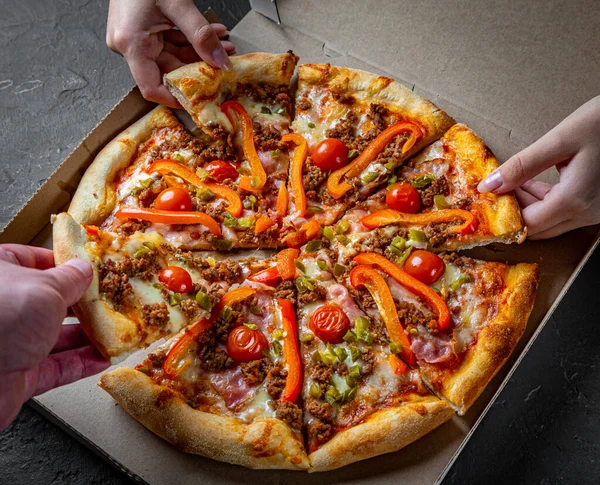 Hands takes a slice of Pizza with Mozzarella cheese, Bolognese sauce, minced meat, pepper, tomato, bacon and vegetables in paper box. Italian pizza on Dark grey black slate background