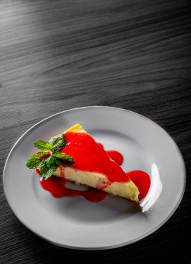 Beyaz tabakta servis edilen çilekli cheesecake siyah ahşap masa arkasında.
