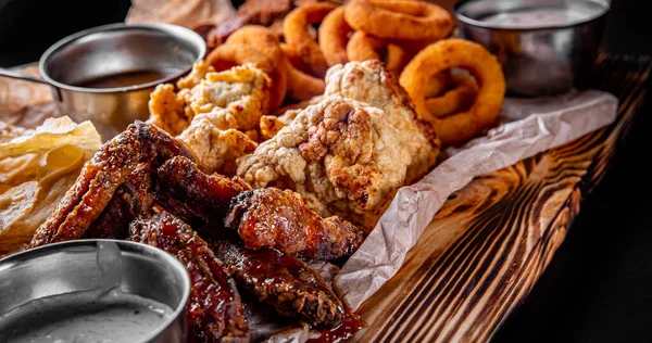 Baharatlı tavuk kanatlı bira tabağı, cips, patates kızartması soğan halkaları, peynir topları, ekmek, ahşap masada tartar sosu.