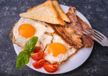Pastırma ve kızarmış yumurta kahvaltı. Gevrek tost dilim dilimlenmiş kiraz domates, fesleğen yaprakları ile beyaz tabakta servis. Karanlık grunge beton tablo arka planı.